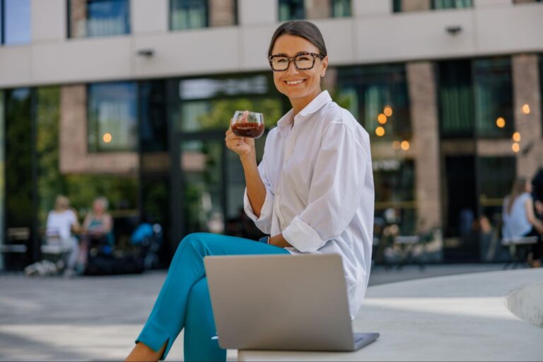 Cheerful woman sipping coffee while working on a laptop outdoors.
